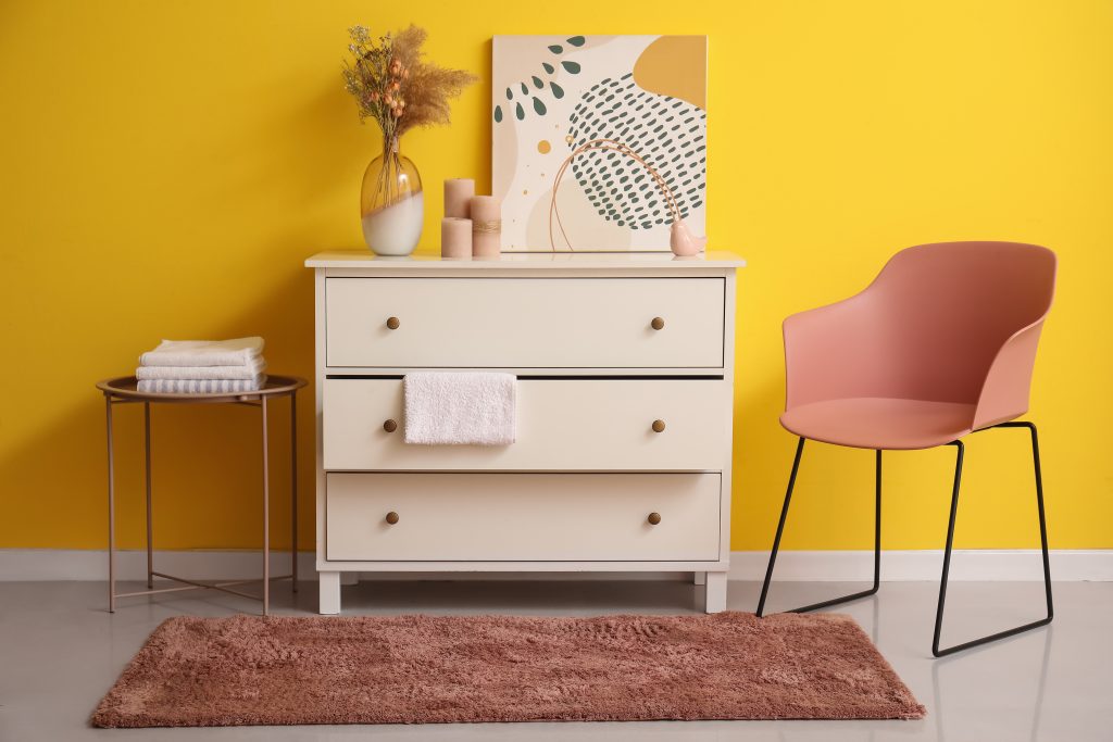 White chest of drawers with decor and stylish chair near yellow wall