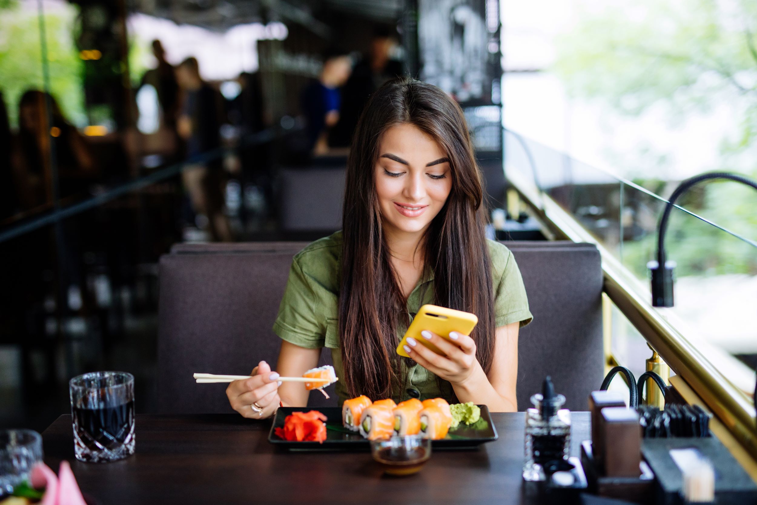 A woman eating sushi in a restaurant and browsing on her phone.